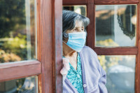 an elderly woman stands in a doorway looking out and is wearing a mask
