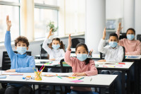 Schoolchildren raising hands at classroom, wearing medical masks stock photo