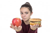 A child holds up an apple and a hamburger with a quizzical expression on her face