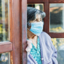 an elderly woman stands in a doorway looking out and is wearing a mask