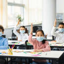 Schoolchildren raising hands at classroom, wearing medical masks stock photo