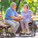 An elderly couple sit in a park, one person in a wheelchair, the other on a bench
