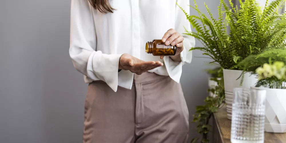 A person in a white shirt and beige pants pours pills from a brown bottle into their hand. Nearby, a potted fern and a glass of water are on a wooden table.