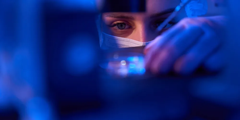 Scientist wearing a mask and hairnet closely examines a sample with a pipette under blue lab lighting.