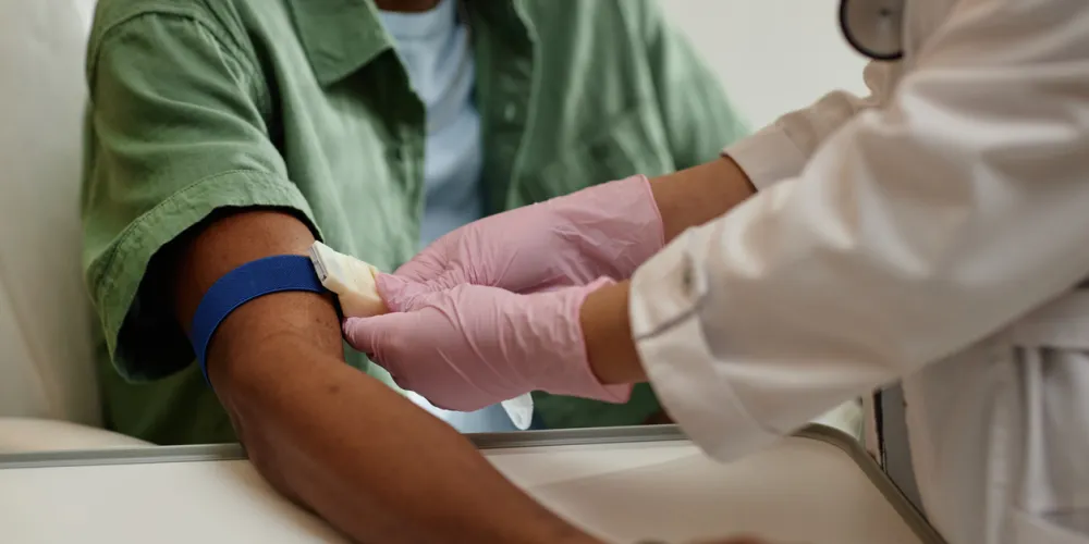 A healthcare professional in a white coat and pink gloves prepares a patient's arm for a blood draw, using a blue tourniquet. The scene is calm and clinical.