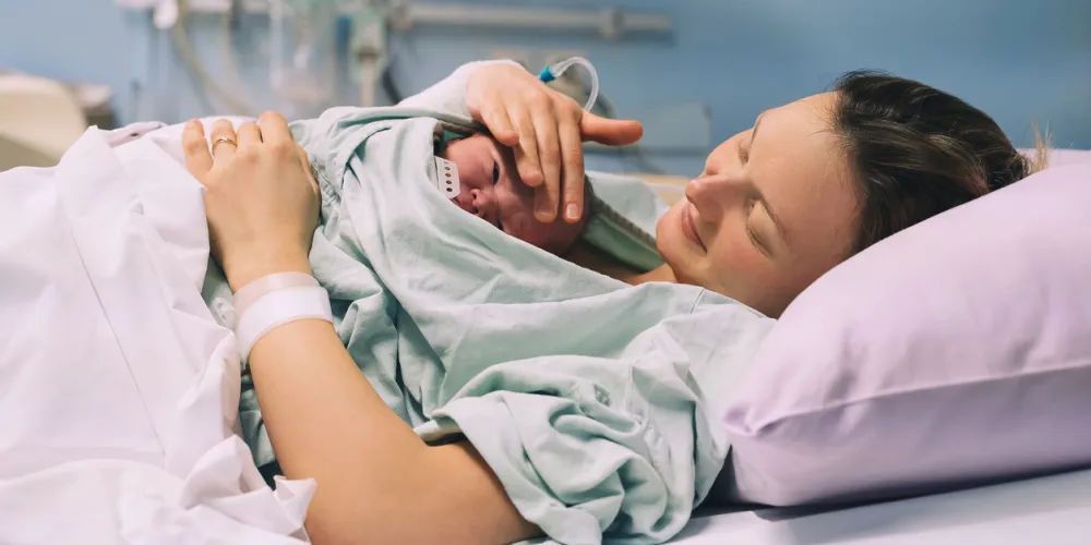 A mother lies in a hospital bed, cradling her newborn wrapped in a green blanket. She smiles, conveying warmth and tenderness in a clinical setting.