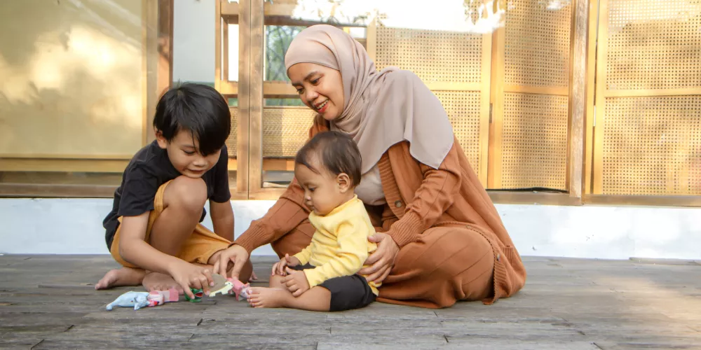 Happy Asian Muslim mom in hijab playing with her two kids on a wooden deck outside, bonding and smiling in warm light