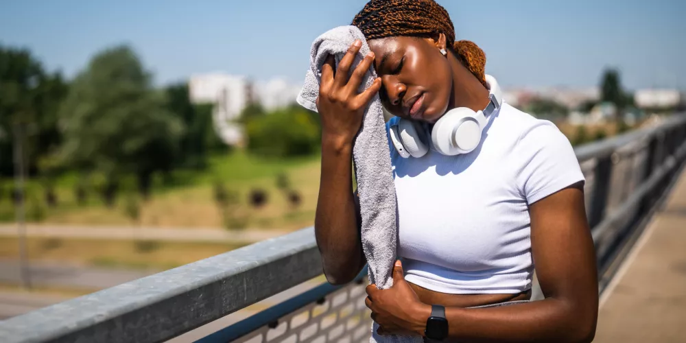 Woman in workout attire rests on a sunny bridge, holding a towel to her forehead. She wears headphones, conveying a sense of exertion and relaxation.