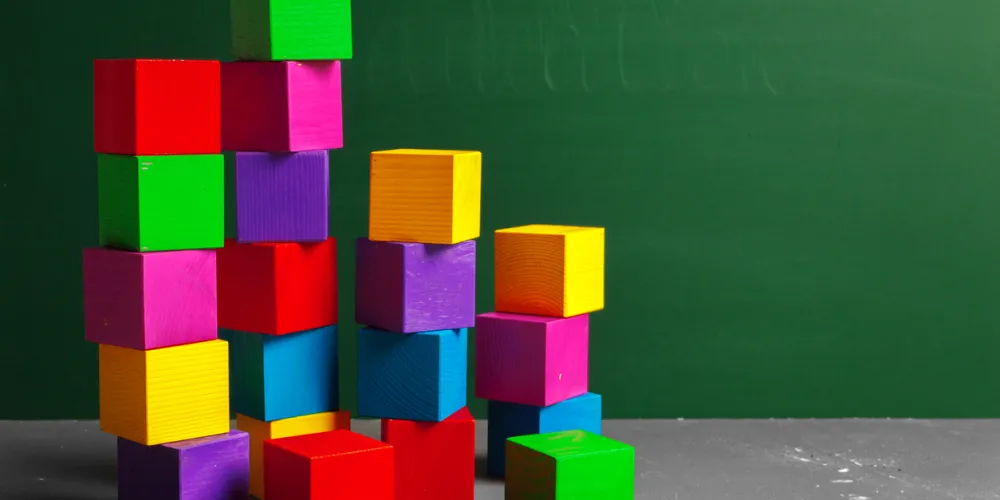 Brightly colored wooden blocks are stacked on a gray surface in a classroom with a green chalkboard
