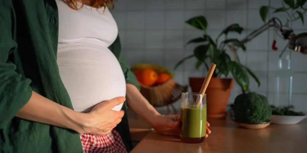 Pregnant woman holding her belly, standing in a kitchen and holding a vibrant green smoothie 