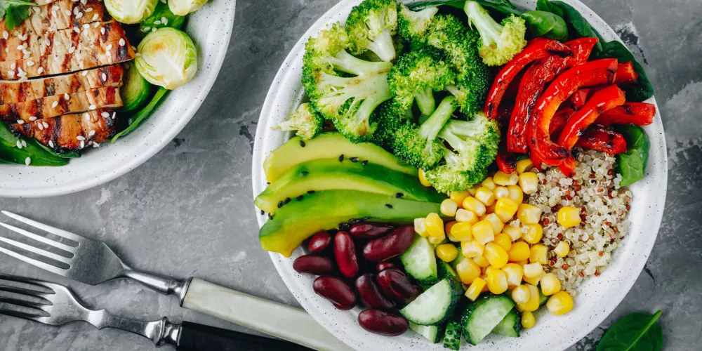 A vibrant salad bowl featuring broccoli, red peppers, avocado, corn, kidney beans, cucumber, and quinoa, with a grilled chicken salad beside it on a gray surface.